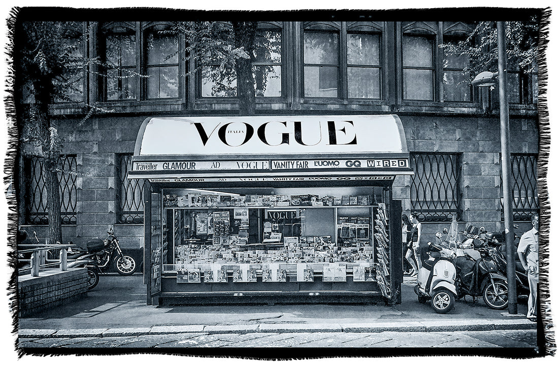 A SUZI ROHER “Lily, Vogue Italia B&W” artwork depicts a city street newsstand with a prominent VOGUE sign, selling magazines and newspapers. Nearby, scooters and a motorcycle are parked—all rendered in striking black and white.