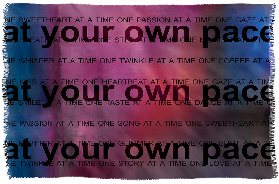 A woven blanket from SUZI ROHER, named “Lily, Your Own Pace,” featuring a pink and blue gradient background. Bold black text declares “at your own pace” in large font, accompanied by smaller phrases such as “one kiss at a time” and “one passion at a time.”.