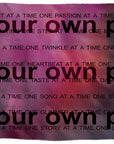 A woven blanket from SUZI ROHER, named “Lily, Your Own Pace,” featuring a pink and blue gradient background. Bold black text declares “at your own pace” in large font, accompanied by smaller phrases such as “one kiss at a time” and “one passion at a time.”.