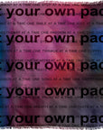 The “Your Own Pace” woven blanket by SUZI ROHER features the phrase “at your own pace” in bold black letters across a vibrant, colorful background adorned with smaller motivational messages.