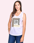 A woman with long brown hair smiles at the camera, wearing SUZI ROHER's The Lola—a white sleeveless top with a geometric wave-riding sketch—paired with blue jeans, standing against a pale pink background.
