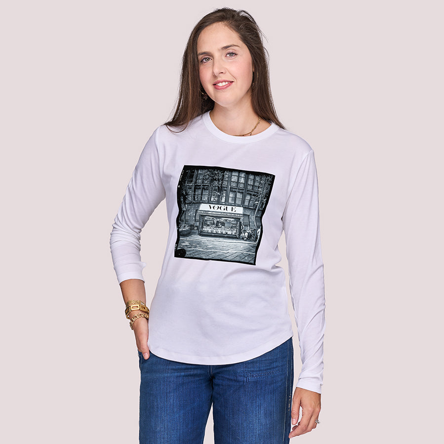 A woman with long brown hair models The Catherine by SUZI ROHER—a white long-sleeve shirt with a black-and-white “APPLE” storefront graphic—smiling brightly against a plain light background.