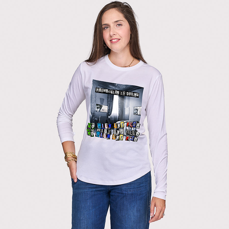 A smiling woman with long brown hair wears The Catherine white slim fit shirt by SUZI ROHER, featuring a colorful graphic and text, paired with blue jeans, standing against a plain light background with one hand in her pocket.