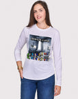 A smiling woman with long brown hair wears The Catherine white slim fit shirt by SUZI ROHER, featuring a colorful graphic and text, paired with blue jeans, standing against a plain light background with one hand in her pocket.