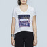 A woman with dark hair and bright lipstick wears The Velvet T-shirt by SUZI ROHER, featuring a white V-neckline and a storefront print labeled VOGUE with "UNPLUGGED" in bold above. She accessorizes with a large bracelet.