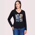 A woman with long brown hair smiles with one hand in her jeans pocket, wearing SUZI ROHER's "The Victoria"—a black long-sleeve V-neck shirt featuring a colorful graphic and supportive words, against a plain light background.