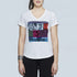A woman wears The Viv by SUZI ROHER, a slim fit white 100% cotton V-neck tee with a colorful patchwork graphic featuring a 7UP logo, $5 sign, and "captcha" against a plain light background.