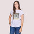 A woman with long brown hair, wearing The Karlie graphic t-shirt by SUZI ROHER and blue jeans, smiles in a relaxed pose against a plain light background, accessorized with gold bracelets and hoop earrings.