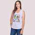 A woman with long brown hair smiles at the camera in The Lola, a sleeveless white top by SUZI ROHER featuring a colorful graphic print, paired with blue jeans and standing gracefully against a plain light background.