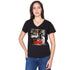 A woman with long brown hair and layered necklaces stands with one hand on her hip, wearing SUZI ROHER’s The Viv—a black V-neck T-shirt with a colorful graphic and the word "ensemble"—paired with blue jeans.