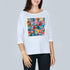 A woman models The Bailey by SUZI ROHER—a white, oversized boxy tee featuring a vibrant geometric patchwork of triangles and unique designs—standing against a plain light gray background.