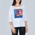 A woman wears The Bailey tee by SUZI ROHER, featuring a white boxy cut and a colorful book spine print with titles like “MONA LISA” and “AMOUR,” posed against a plain gray background.