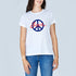 A smiling woman with long brown hair wears The Niki by SUZI ROHER, a 100% cotton white tee featuring a blue peace symbol and “Love” in red script, standing against a light gray background.